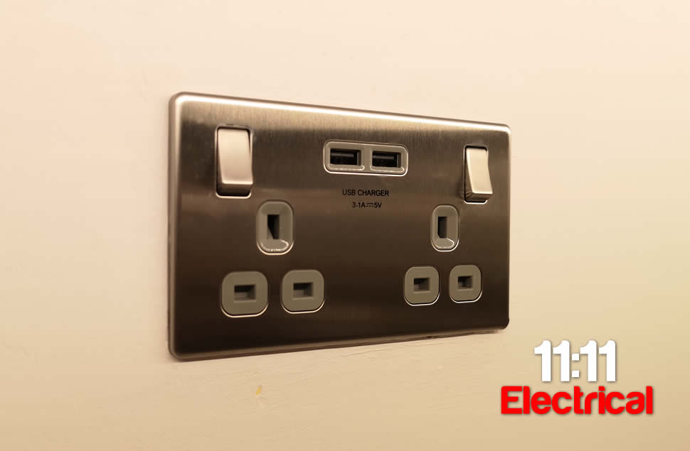 Brushed steel UK double socket with USB outlets installed on warmly lit plasterboard grey wall by 11:11 Electrical in Bournemouth